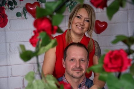 Paar im Vordergrund umgeben von roten Rosen und Herzen, lächelnd in die Kamera.