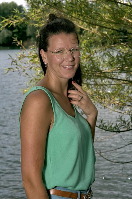 Frau mit Brille, grünem Oberteil und lächelndem Gesicht vor einem See.