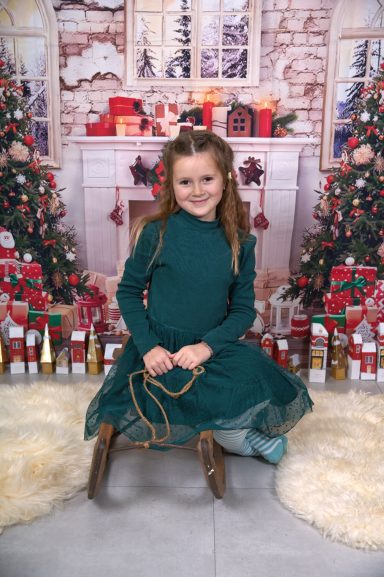 Mädchen in einem grünen Kleid sitzt vor einem festlich dekorierten Hintergrund mit Weihnachtsbaum.