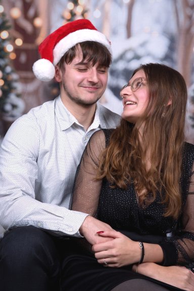 Paar lächelt sich an, festlich gekleidet, mit Weihnachtsdekoration im Hintergrund.