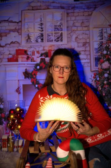 Frau mit Brille trägt einen Weihnachtspullover und hält eine leuchtende Dekoration.