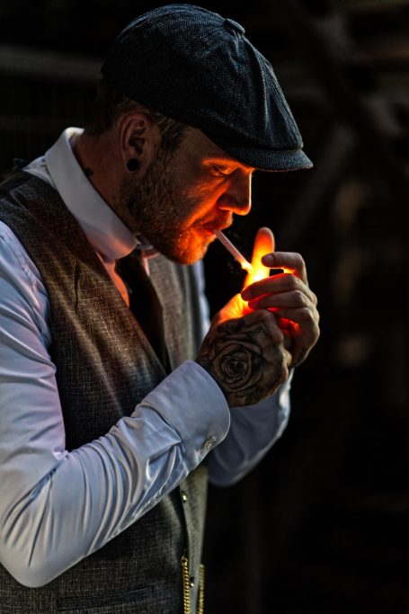 Ein Mann mit Hut und Weste zündet eine Zigarette an. Tätowierungen sind sichtbar.