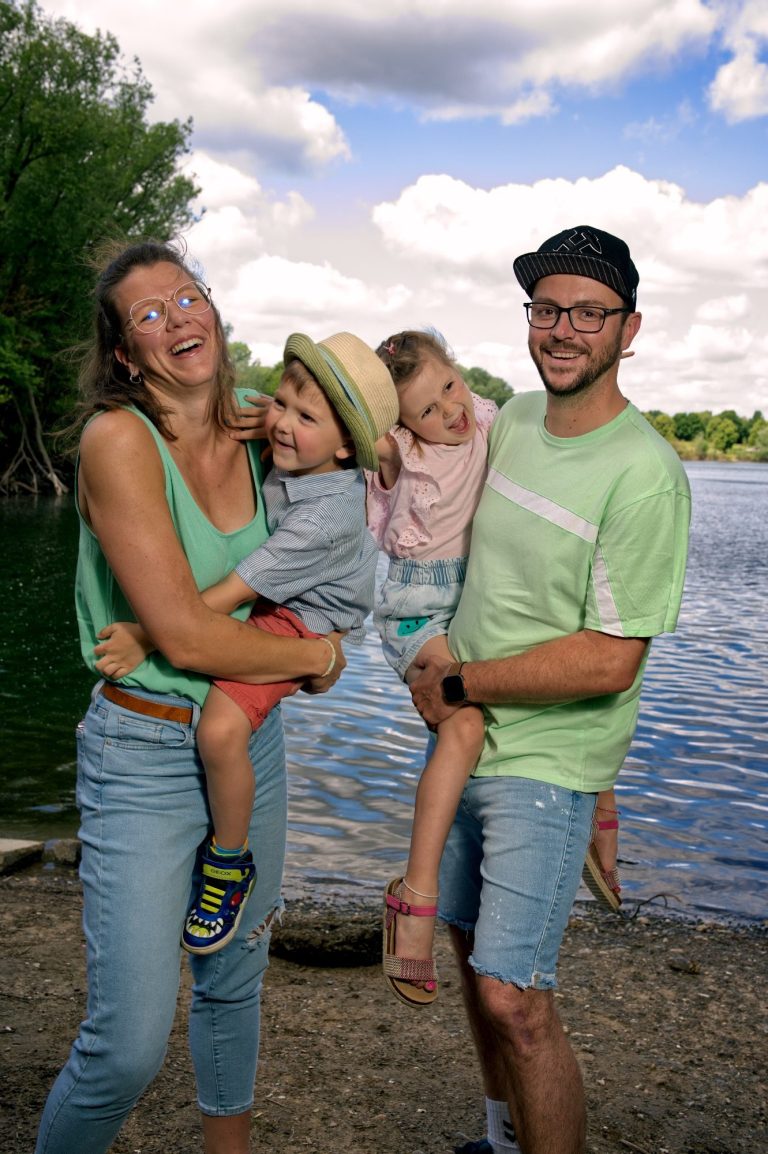 Familie posiert am Wasser, zwei Kinder und Erwachsene lächeln fröhlich.