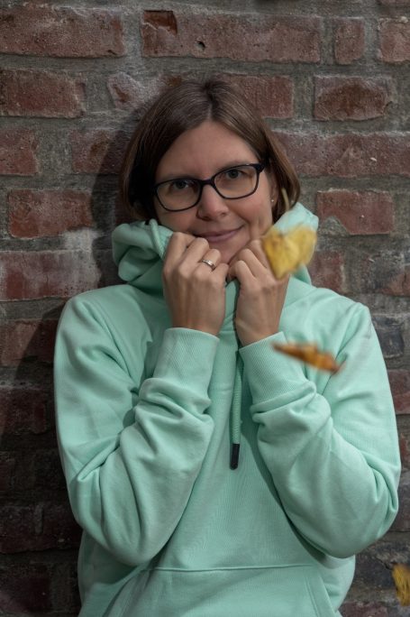 Junge Frau in mintgrünem Hoodie mit Brille, lächelt schüchtern vor einer Backsteinwand.