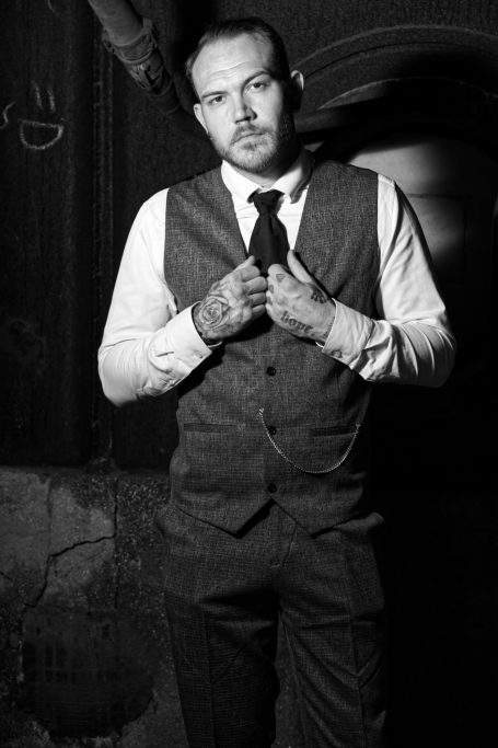 Mann in Anzug mit Krawatte, ernstes Gesicht, stehend vor dunklem Hintergrund.