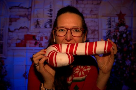 Frau mit Brille hält ein rot-weiß gestreiftes Objekt vor einem festlichen Hintergrund.