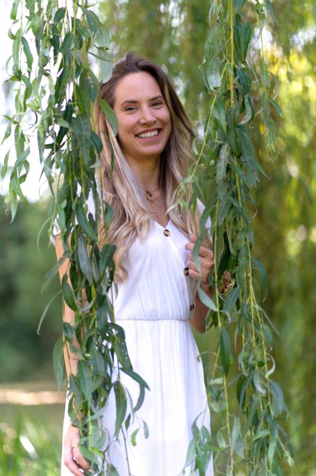Junge Frau in einem weißen Kleid lächelt hinter hängenden Zweigen eines Baumes.