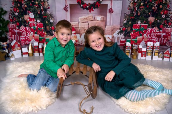 Zwei Kinder, ein Junge in grünem Pullover und ein Mädchen in grünem Kleid, lächeln vor Weihnachtsdekoration.