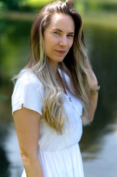 Frau mit langen, blonden Haaren in einem weißen Kleid, steht am Wasser.