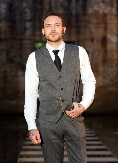 Mann in Anzug mit Weste und Krawatte steht vor einer rustikalen Hintergrundkulisse.