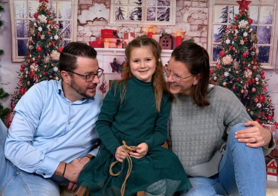 Ein Kind in einem grünen Kleid sitzt zwischen zwei Erwachsenen vor Weihnachtsdekoration.