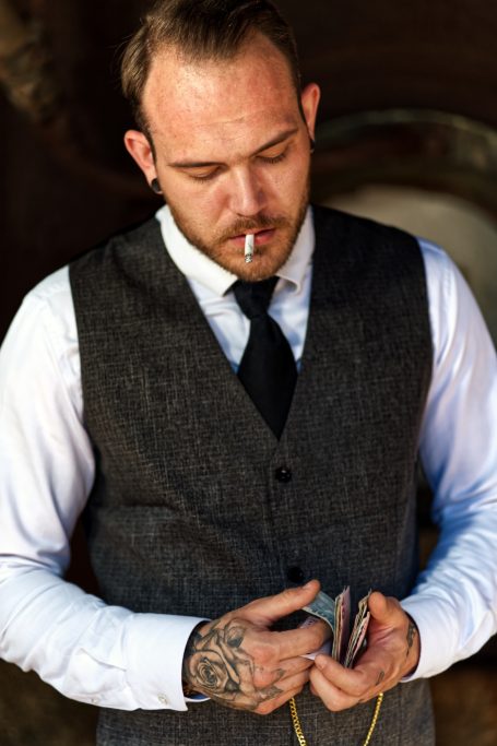 Mann in Anzug mit Zigarette, hält eine Geldbörse und zeigt sichtbare Tätowierungen.