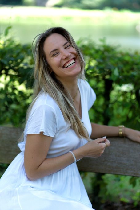 Lachende Frau mit langen Haaren, die auf einer Bank im Freien sitzt, umgeben von Pflanzen.