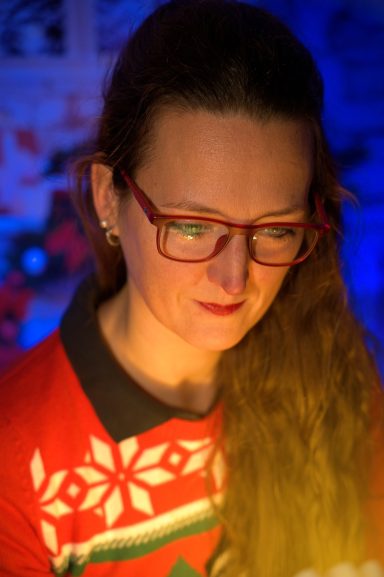 Frau mit langen, braunen Haaren und Brille, trägt ein weihnachtliches Sweater.